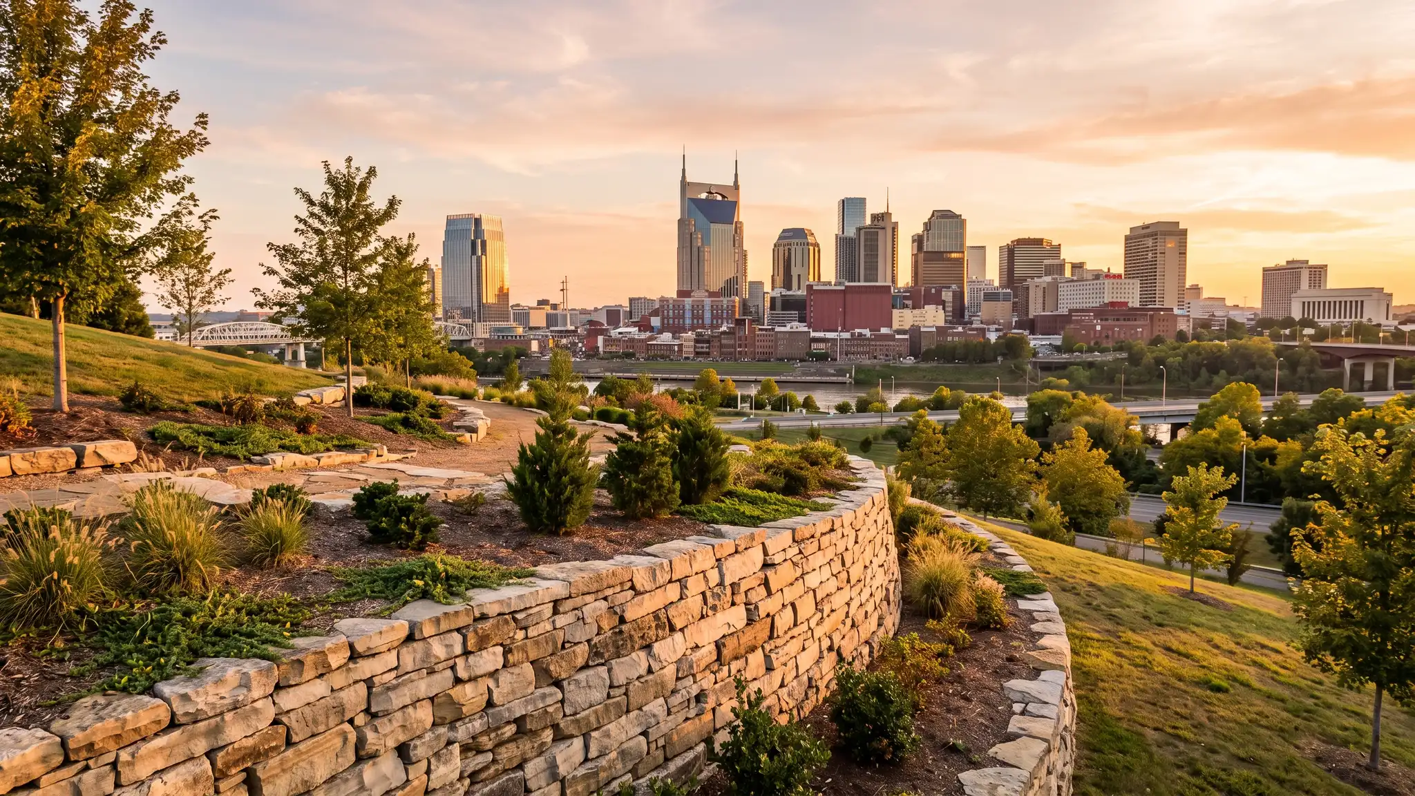 A golden hour view of the Nashville skyline from a landscaped hillside with a stone retaining wall in the foreground